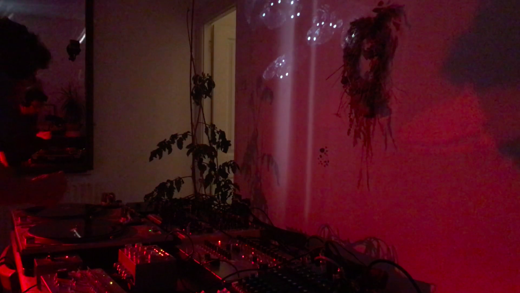 room lit with low pink light showing musician in front of synthesisers and turntables