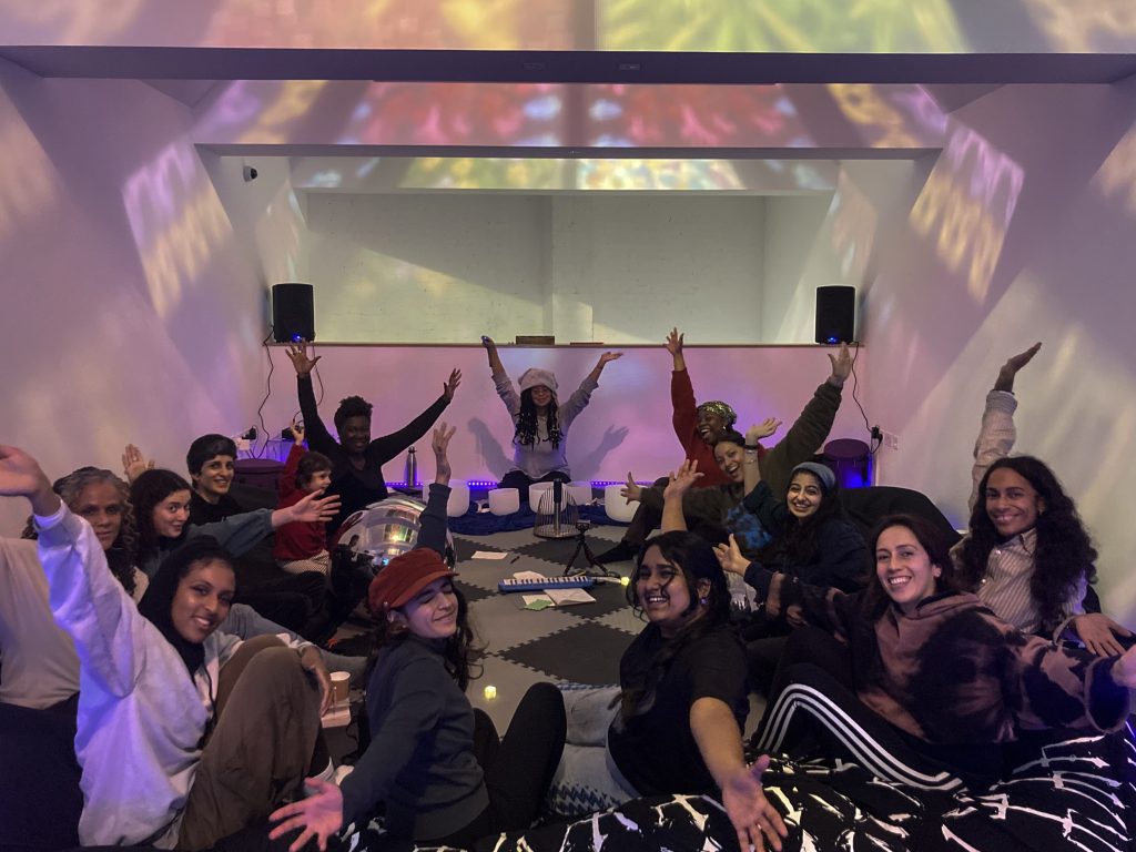 A group of queer non-binary and women of colour are sat on the floor in a circle. They turn to smile and wave at the camera.