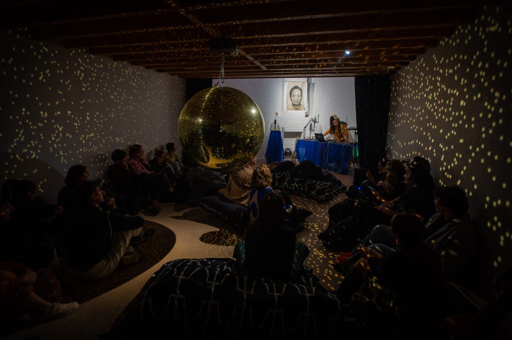 Documentation of a performance by HCJ within the exhibition. The mirror ball from 'Owed to Chiron the Wounded Healer' is shown at the front where people are sat watching the performance.